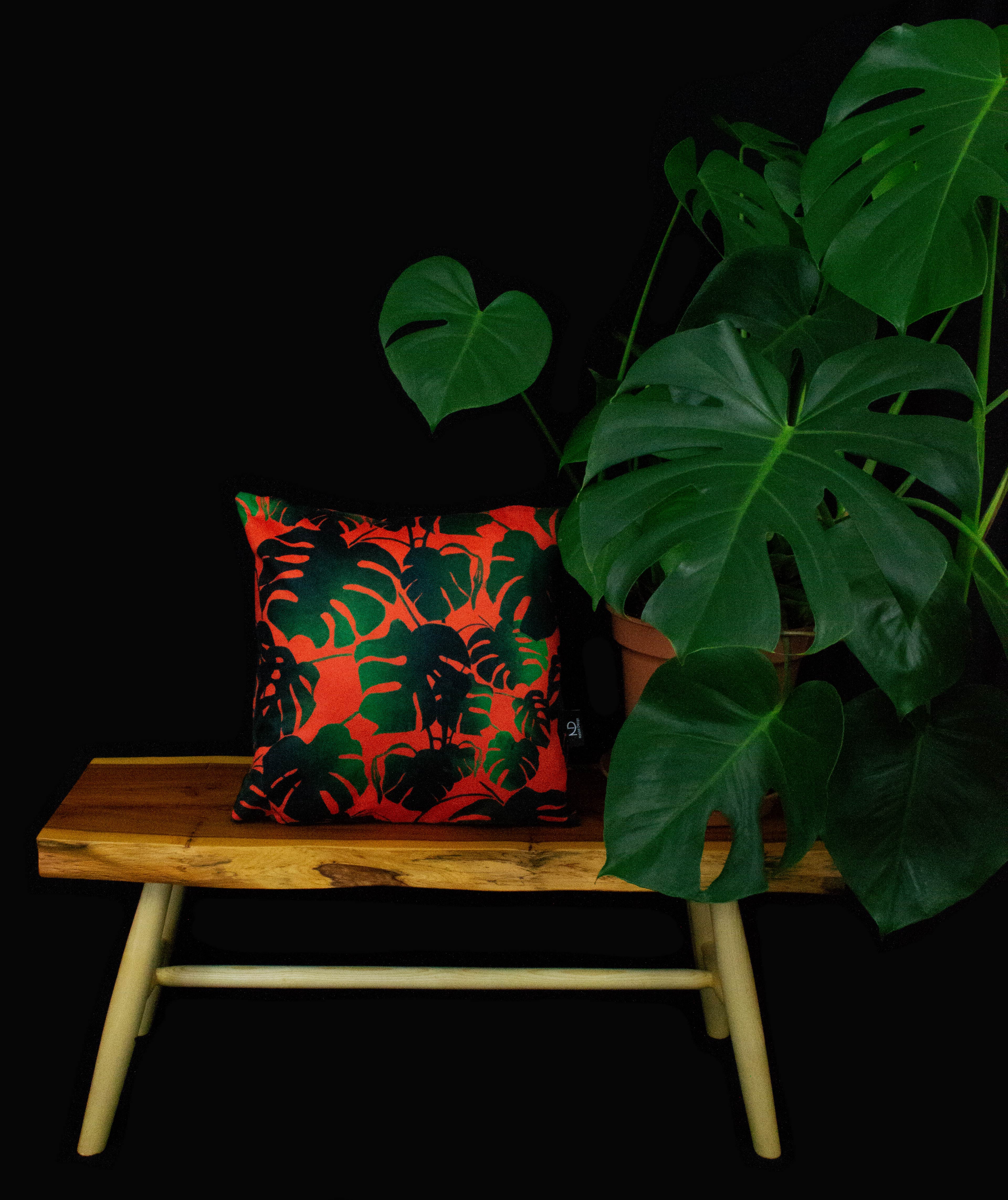 Cheese plant cushion with green printed leaves ontop of a orange backgroun displayed ontop of wooden table.